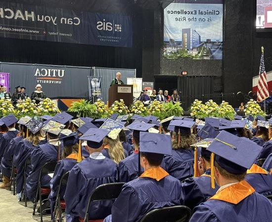 Members of the Class of 2024, 戴着帽子，穿着长袍, sit in the Utica Memorial Auditorium as President Todd Pfannestiel speaks from a podium.